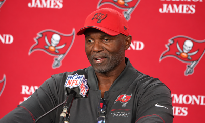 Todd Bowles wears a red Buccaneers cap and dark team shirt while speaking into an NFL microphone against a red Buccaneers and Raymond James backdrop.