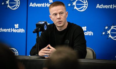 Orlando Magic coach Sean Sweeney speaks into a microphone at a press table against a blue backdrop featuring Magic and AdventHealth logos.